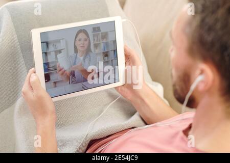 Young sick man with digital tablet sitting on couch under blanket while having rest and listening to online doctor advice about medicine Stock Photo