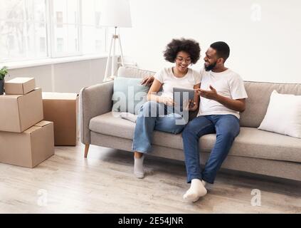 Happy Black Spouses Using Tablet Sitting Among Moving Boxes Indoor Stock Photo