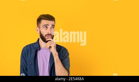 Dreamy Caucasian guy thinking over something, touching his chin on orange background, banner design with copy space Stock Photo