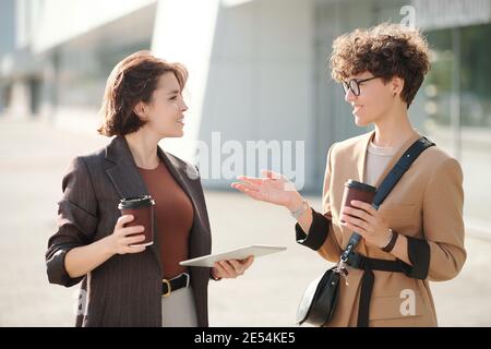 Two young female colleagues with tablet discussing working points in urban environment while having drinks against business center Stock Photo