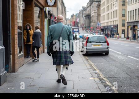 Edinburgh Scotland - October 2019 Scottish man walking along high street in tartan kilt with bag and green jacket through city centre from behind look Stock Photo