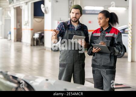 Young confident repairman in uniform describing his female colleague characteristics of one of cars while using data from digital tablet Stock Photo