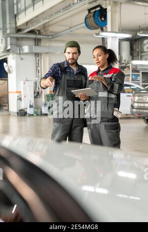 Young repairman and his female colleague in uniform discussing characteristics of car in workshop while using data from digital tablet Stock Photo