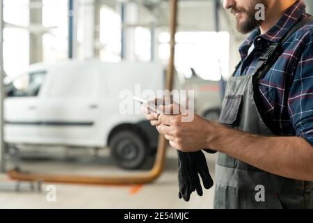 Side view of young bearded worker or repairman of car maintenance service using touchpad while standing in front of camera in workshop Stock Photo