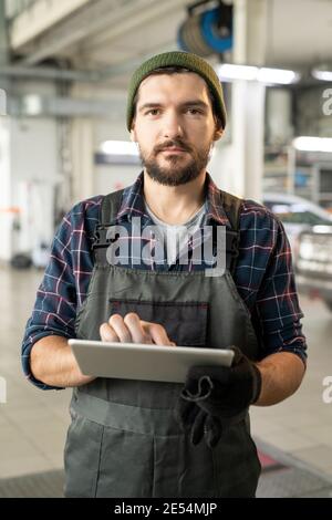 Young serious male worker of car service using digital tablet while answering online questions of clients in working environment Stock Photo