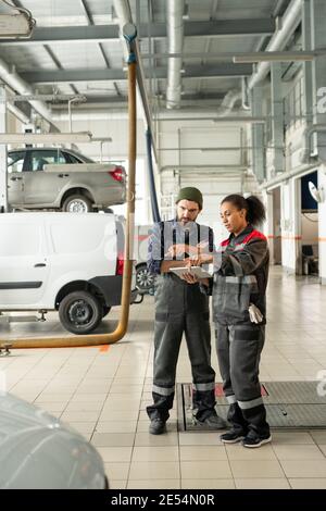 Young confident technicians in workwear pointing at one of cars in workshop while discussing its technical characteristics before repairing it Stock Photo