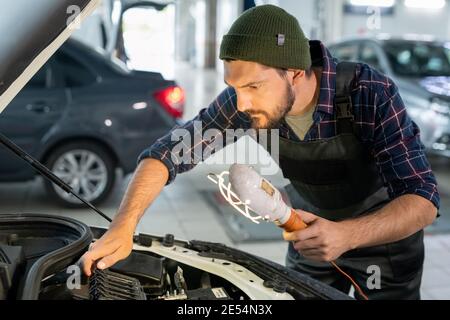 Young serious professional repairman in workwear holding lamp and bending over open hood while repairing broken car engine Stock Photo