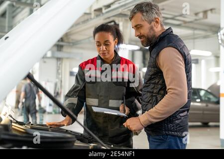 Young female technician of car repair service looking at open engine compartment while describing problem to male client in workshop Stock Photo