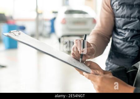 Hand of female technician of automobile repair service holding document while male client with pen putting his signature after repairing car Stock Photo