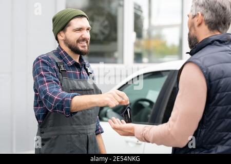 Happy young repairman giving alarm system remote control to male client after repairing automobile against car and interior of workshop Stock Photo