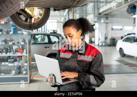 Female worker of car repair service in uniform using laptop in workshop to look for data in online sources, take new orders or consult clients Stock Photo