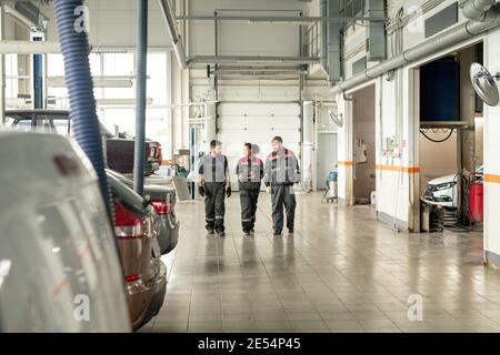 Three intercultural male and female workers of car maintenance center moving along large workshop and discussing working points Stock Photo