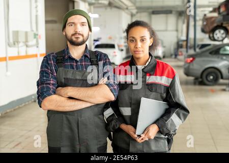 Two young workers of car repair center standing close to each other in front of camera against interior of large workshop with automobiles Stock Photo