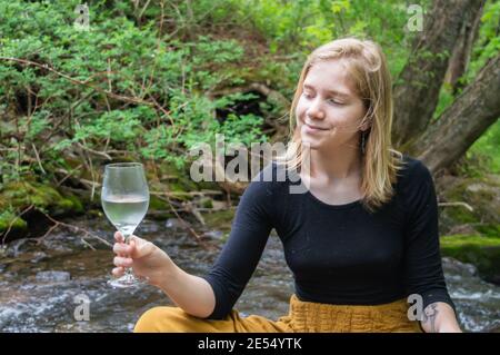 Portrait of a young smiling woman sitting cross legged on a moss covered rock on the shore of a river in the woodland, tasting a glass of white wine. Stock Photo