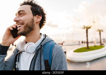Cheerful young guy smiling and talking on mobile phone while walking on promenade Stock Photo