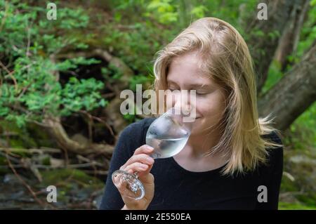 Close up shot of a young woman sitting on the shore of a river in the woodland, tasting a glass of white wine and smelling it's perfume. Stock Photo
