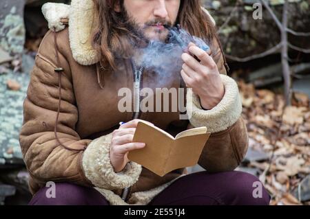 Portrait shot of a young man with long hair and sheepskin coat sitting on a rock, reading a notebook and smoking a pipe during a walk in the country. Stock Photo
