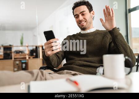 Confident young man manager having a video chat via mobile phone while sitting at the desk in office Stock Photo