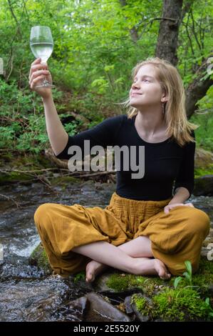 Portrait of a young smiling woman sitting cross legged on a moss covered rock on the shore of a river holding up and looking at a wine glass. Stock Photo