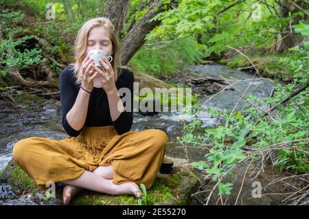 Portrait of a pretty young girl sitting cross legged on a rock on the shore of a river, holding a china tea cup with both hands and drinking