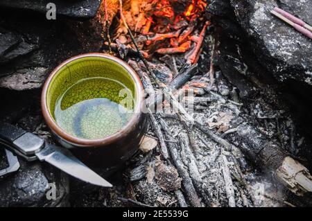 Close up shot from above of a small green china tea cup with water in it and a camping knife at the side warming up next to glowing embers. Stock Photo