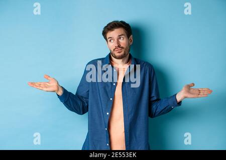 Indecisive young man with beard, look away and shrugging shoulders, know nothing, being clueless, standing on blue background Stock Photo