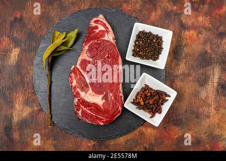 Raw beef ribeye on a round slate plate with glass containers containing peppercorns, coarse salt and cayenne chillies on an old wooden table in brown Stock Photo
