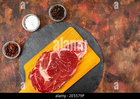 Raw beef ribeye on a round slate plate with glass containers containing peppercorns, coarse salt and cayenne chillies on an old wooden table in brown Stock Photo