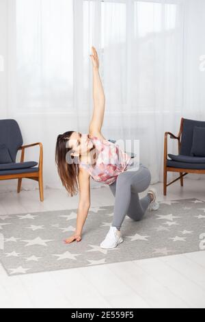 A full-length young fit woman in gray leggings with her legs spread wide makes twists in a home interior with a large white window Stock Photo