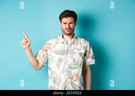 Disappointed tourist cringe and frown, pointing finger aside at empty space, complaining on bad thing, standing in hawaiian shirt on blue background Stock Photo