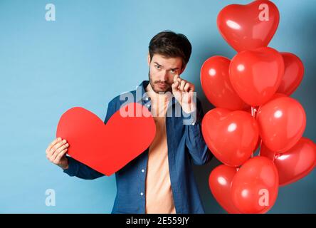 Sad and heartbroken man crying, wiping tears, standing with red heart and balloons, breakup on Valentines day, blue background Stock Photo