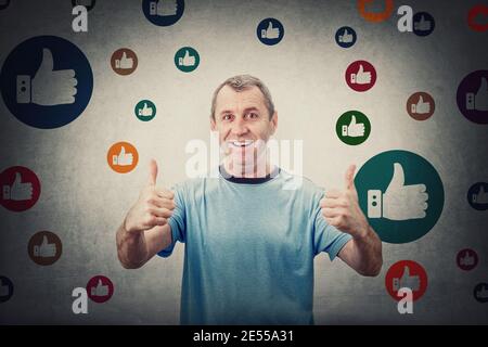 Excited middle aged man, shows thumbs up gesture, smiling contented to camera, surrounded by multicolor like signs. Senior male gives approval symbol, Stock Photo