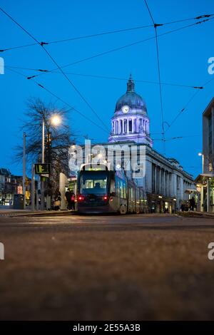 Council house illuminated with tram parked in front lit up night street lamps cables train waiting passengers shoppers to board blue sky Nottingham Stock Photo