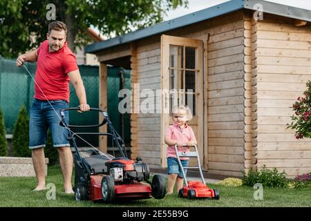 Father and his son mowing the lawn together. Full length of a young boy cutting the grass with his father. Stock Photo