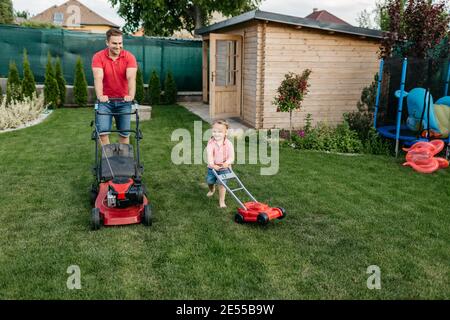 Father and his son mowing the lawn together. Full length of a young boy cutting the grass with his father. Stock Photo