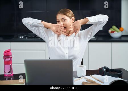 Beautiful young woman sitting at table with opened laptop, stretching and yawning. Pleasant female feeling tired after long working day at home. Stock Photo