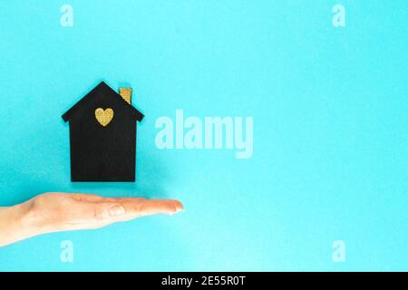A female hand holds a mock-up of a dark house on a blue background. Family, real estate and insurance concept, flat lay, top view. Stock Photo