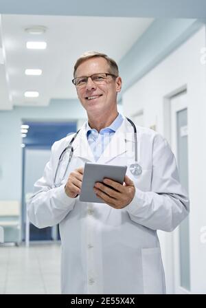 Old male doctor holding digital tablet computer looking at camera in hospital. Stock Photo