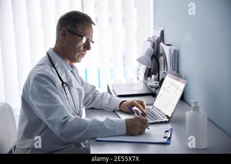Serious mature doctor writing notes, using laptop in medical office. Stock Photo