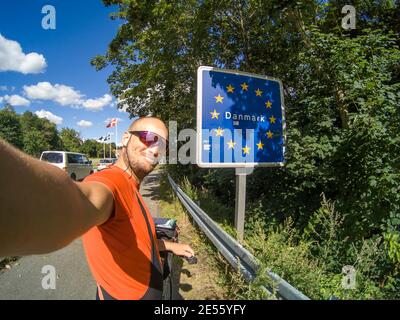 happy handsome attractive tourist on bicycle doing selfie in Flensburg, Germany at Denmark Border with sign saying 'Danmark' (Denmark) Border between Stock Photo