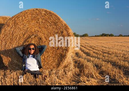 Mixed race African American girl teenager female young woman leaning against hay bale in a field at sunset or sunrise golden evening or morning sunshi Stock Photo