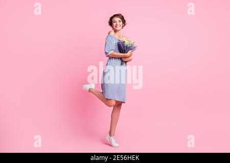 Photo portrait of happy girl holding floral bouquet in two hands standing on one leg isolated on pastel pink colored background Stock Photo