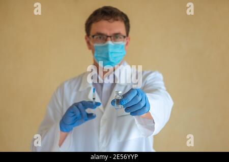 A man doctor in a protective mask and blue latex gloves holds an ampoule with a vaccine and a syringe. Focus on the ampoule Stock Photo