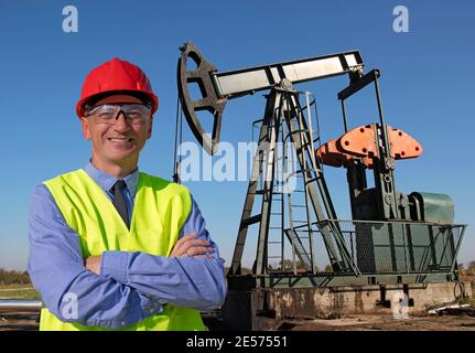 Smiling Engineer in Red Hardhat and Yellow West Standing in front of the Oil Well. Happy Manager Looking at Camera. Portrait of  Oil and Gas Engineer. Stock Photo