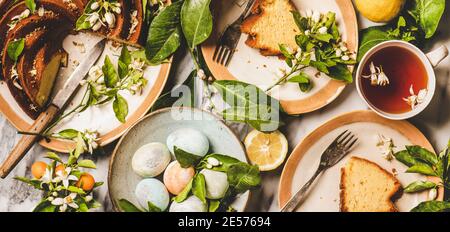 Easter holiday dinner table setting. Flat-lay of colorful Easter Eggs, Lemon cake with citrus tree blossom branches and black tea over white marble table background, top view, wide composition Stock Photo