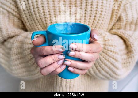 Cold hands. Woman in cozy woolen sweater warming hands with cup of hot tea or coffee. Stock Photo