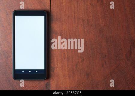Black mobile phone with blank white screen on rustic wooden table. Minimalistic top view of workspace with copy space. Stock Photo