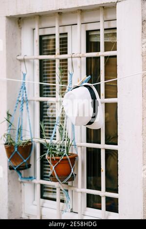 A window with a white metal lattice with flower pots on hooks and a white hat on clothespins. Stock Photo