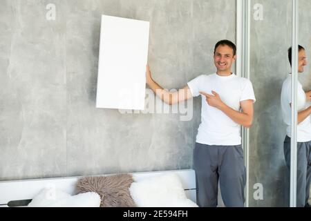 Young man in white t-shirt is holding blank empty canvas. Guy student is smiling. Template for advertising, greeting, invitation. Emotional portrait Stock Photo