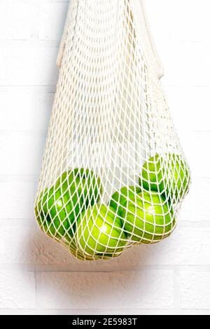Fresh green apples in eco natural reusable cotton mesh bag. Zero waste concept. White brick background.  Stock Photo